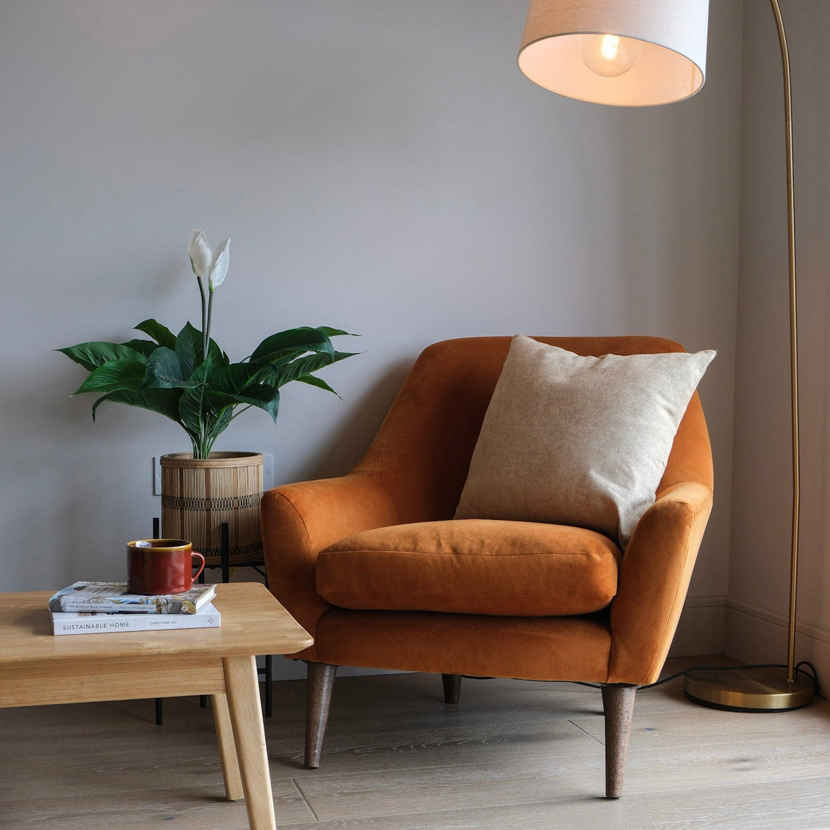 An orange velvet armchair with a beige throw pillow in a minimalist room, next to a wooden coffee table, a potted plant, and a tall floor lamp.