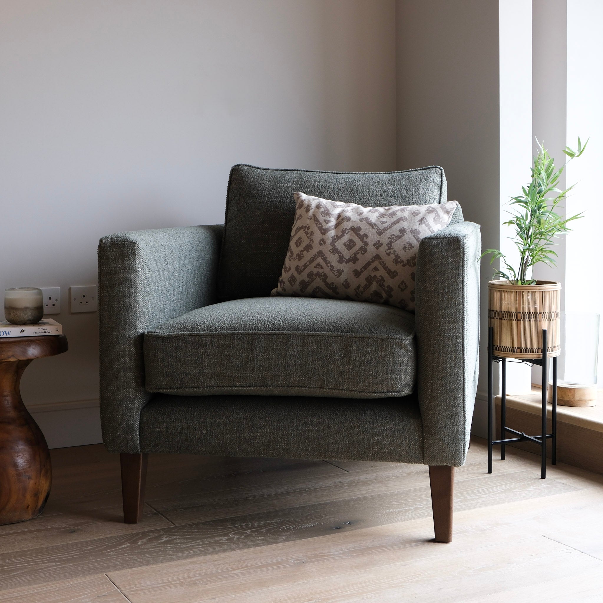 A dark green upholstered armchair with dark wooden legs, styled with a patterned throw pillow, set in a minimalist room with a side table, potted plant, and light wood flooring.