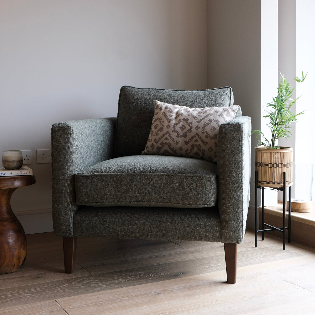 A dark green upholstered armchair with dark wooden legs, styled with a patterned throw pillow, set in a minimalist room with a side table, potted plant, and light wood flooring.
