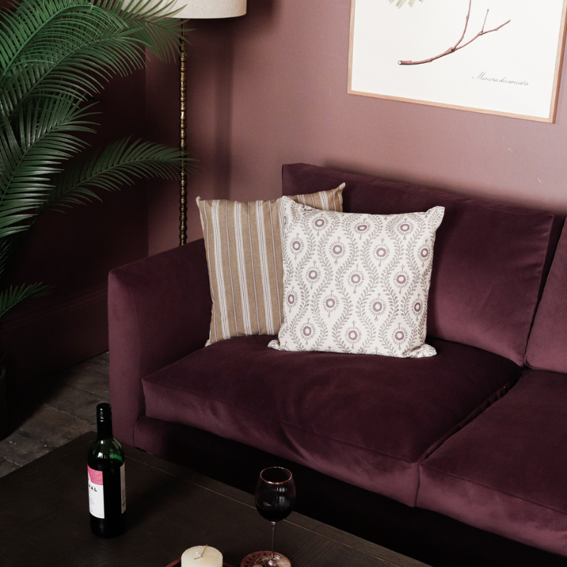 Rum Raisin Purple/Red sofa with patterned cushions in a room with a artificial plant and framed botanical art print on the wall.