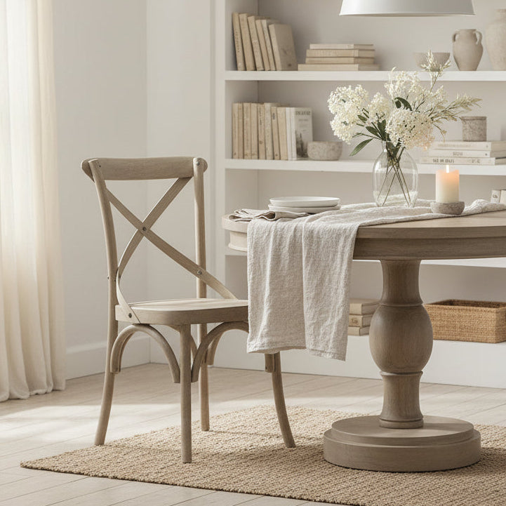 A round light wood dining table with a pedestal base and a matching wooden chair, set in a warehouse or showroom surrounded by stacked furniture pieces wrapped in protective material.