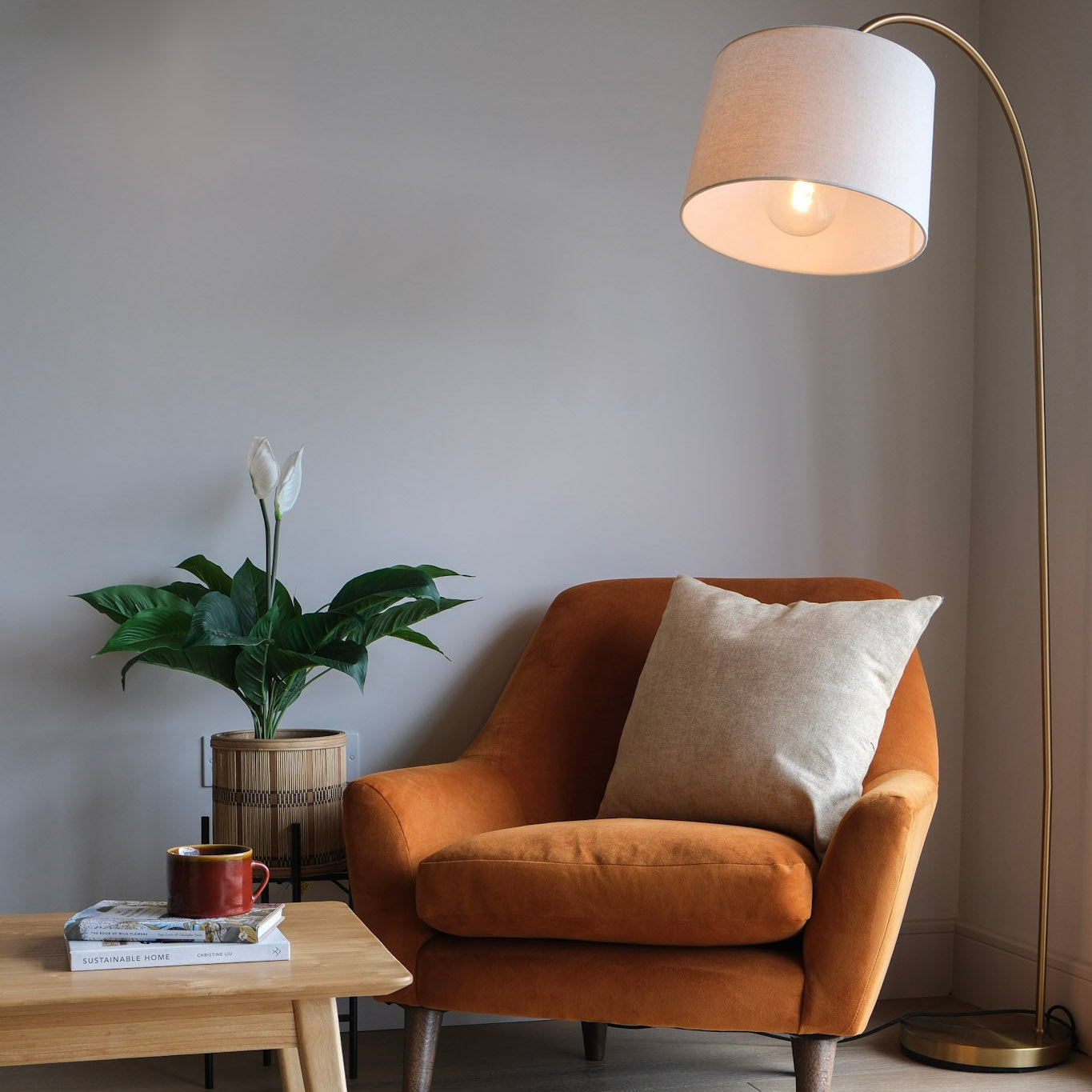 An orange velvet armchair with a beige throw pillow in a minimalist room, next to a wooden coffee table, a potted plant, and a tall floor lamp.