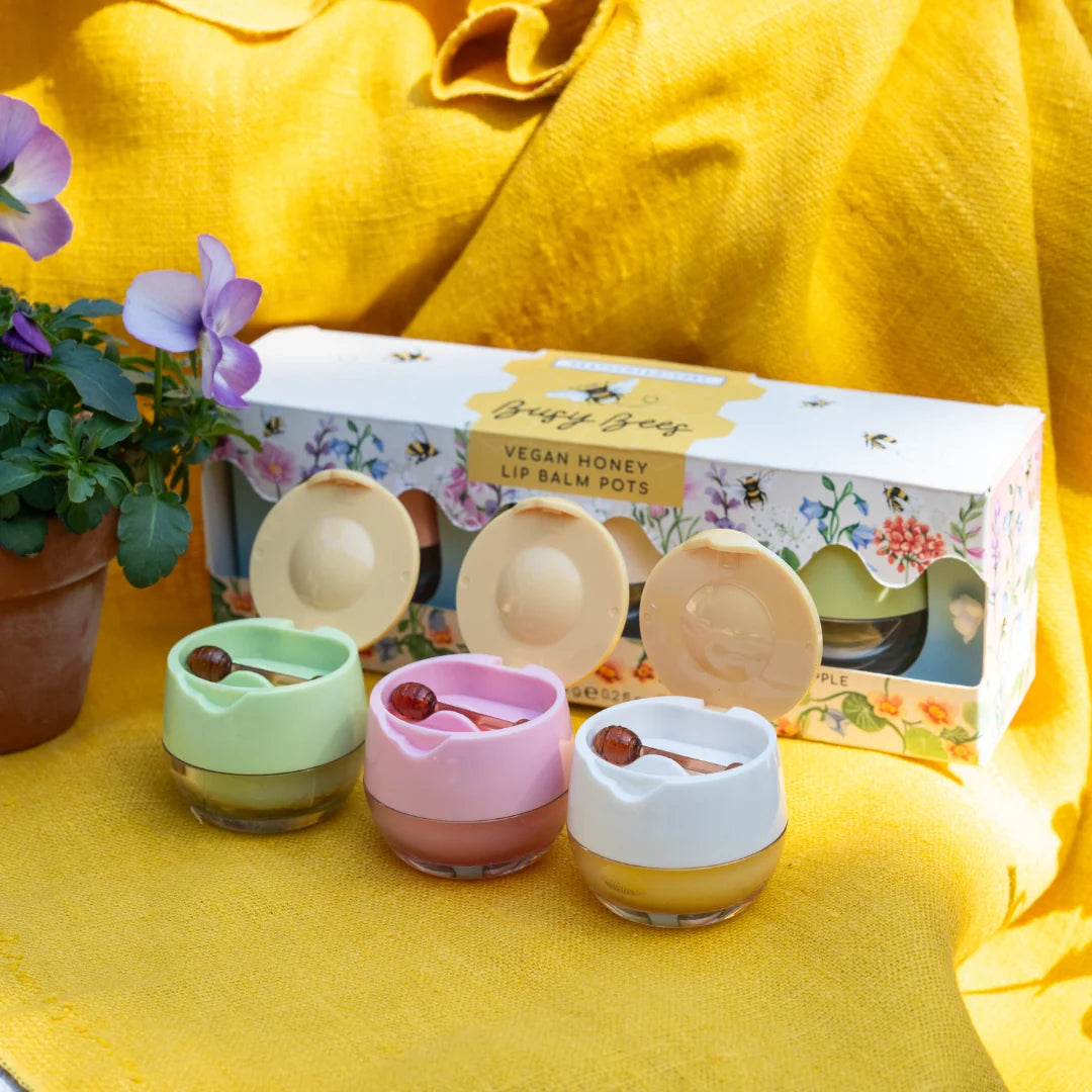 Three small vegan honey lip balm pots in pastel green, pink, and white, arranged in front of their floral product box on a bright yellow fabric backdrop, with a potted plant at the side.