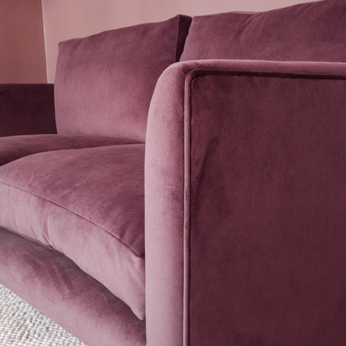 A close-up of a dark purple/red mauve velvet sofa with a dark wood leg, resting on a textured cream woven rug