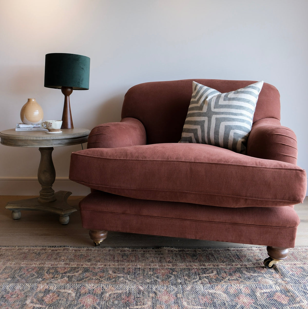 A dark red/brown cotton linen armchair with a matching ottoman, styled in a living room setting next to a wooden side table with a green lamp and a patterned throw pillow.