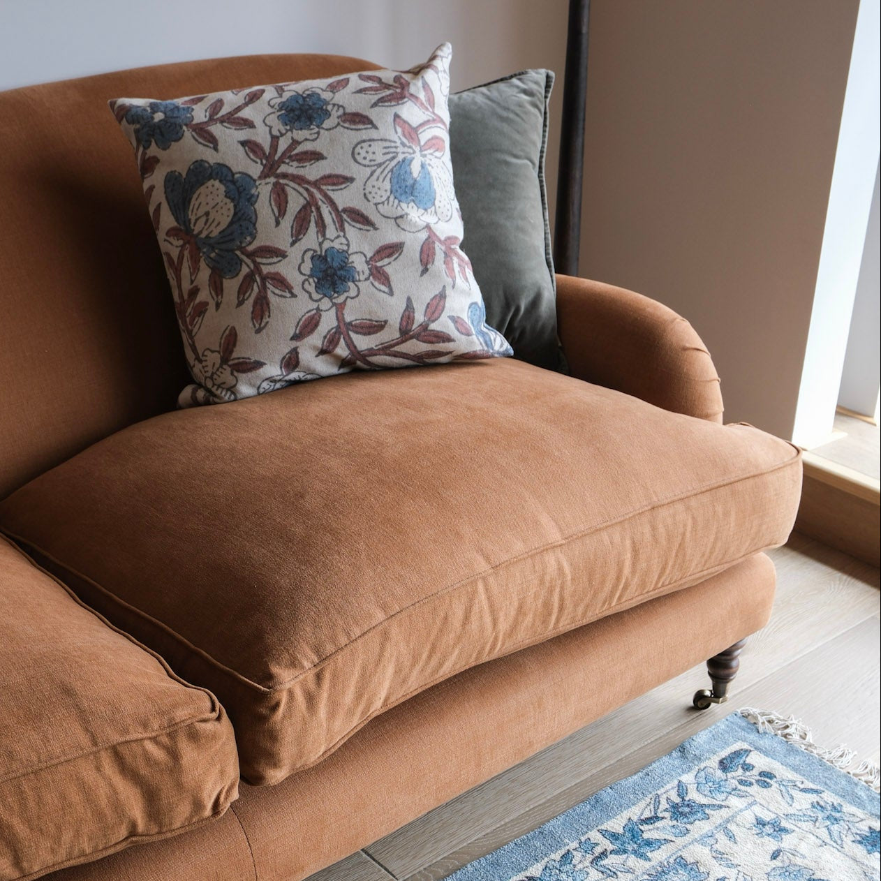 A warm rust-brown velvet loveseat sofa in a styled living room setting, featuring a floral throw pillow and a grey accent pillow, with a wooden coffee table and blue patterned rug in the foreground.