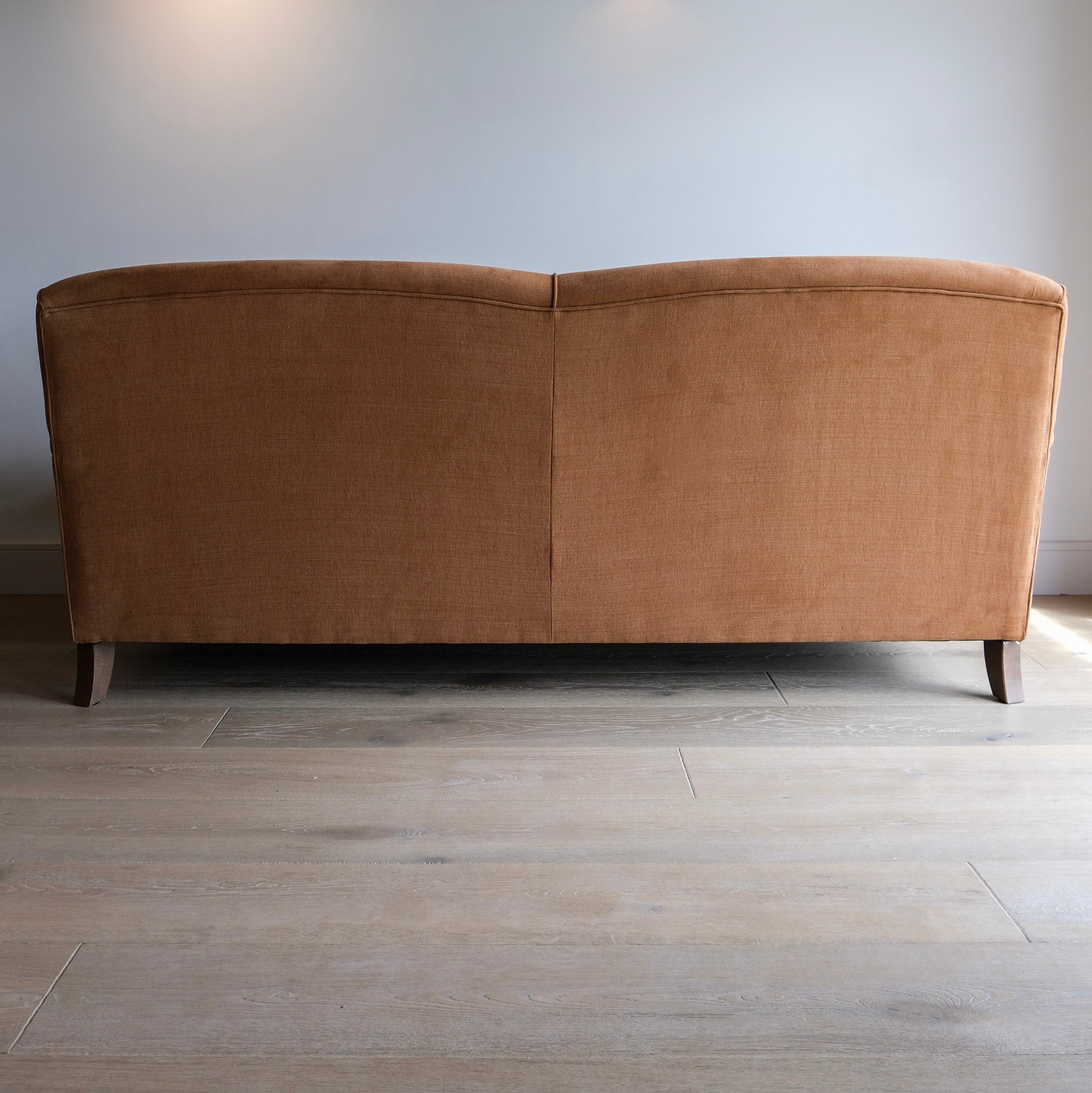 A chocolate brown upholstered sofa shown from the back, positioned against a light gray wall on a light wood floor.