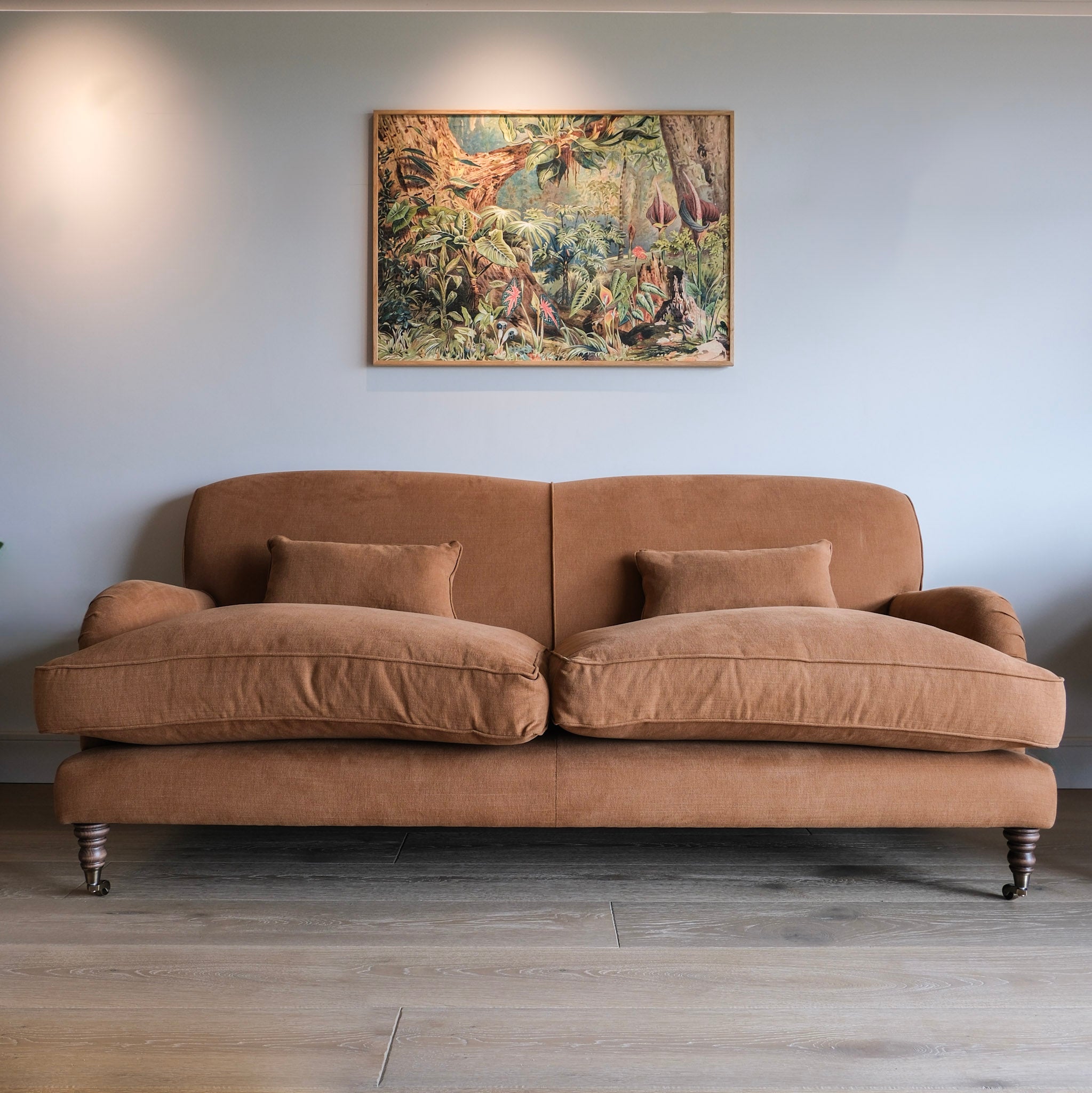 A two-seater upholstered sofa in a warm chocolate brown fabric, positioned against a light gray wall with a framed jungle painting hanging above it. The sofa features two matching throw pillows and dark wooden legs.