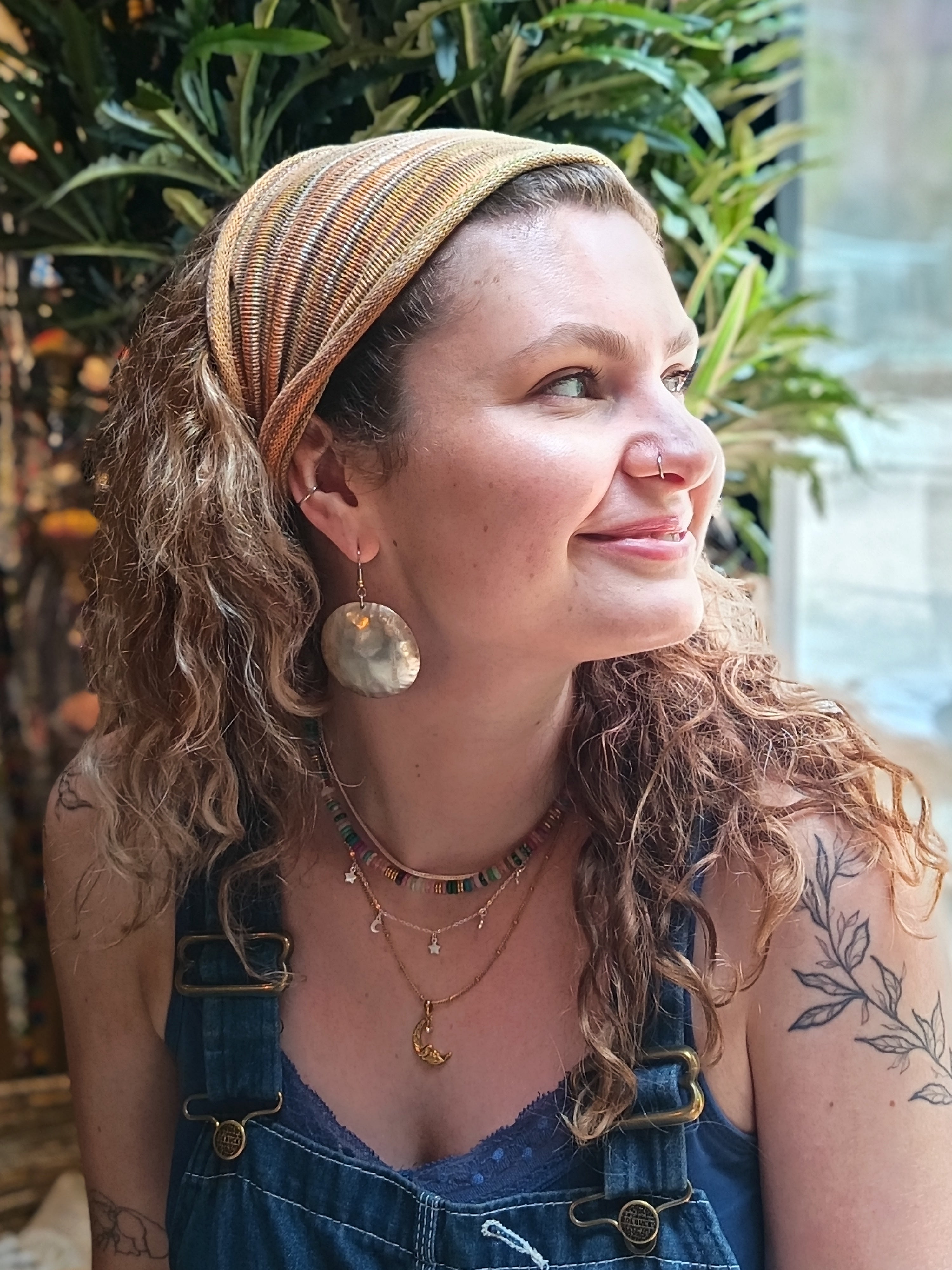 Woman with curly hair wearing a headscarf and denim overalls, sitting indoors with plants in the background.