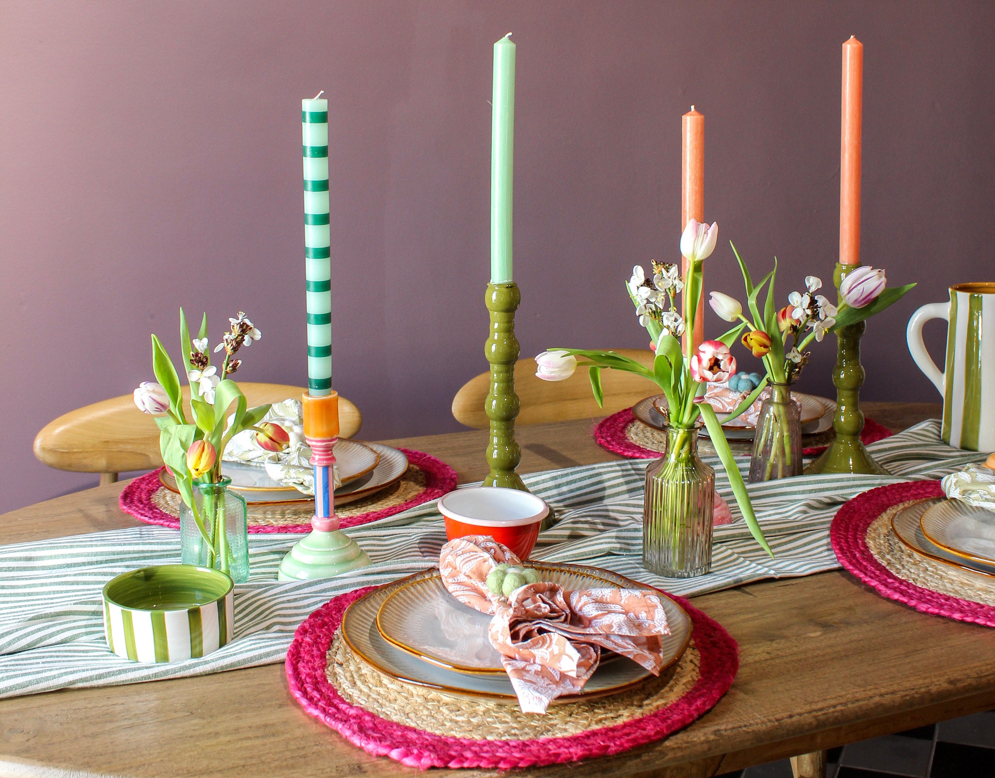Dining table set with colourful candles, flowers, and place settings against a purple wall. 