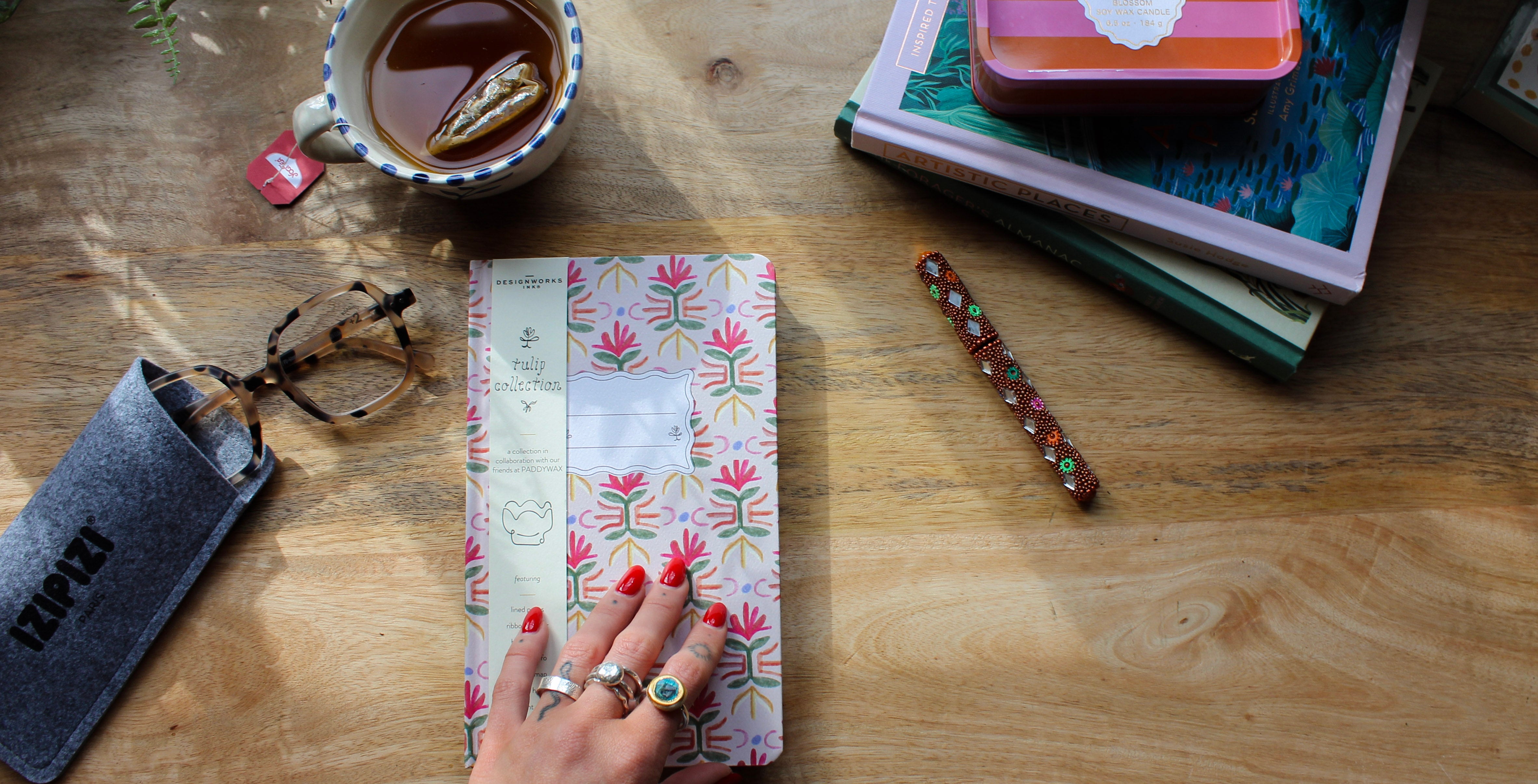 Hand holding a floral notebook on a wooden desk surface with a cup of tea and books and Izipizi sunglasses