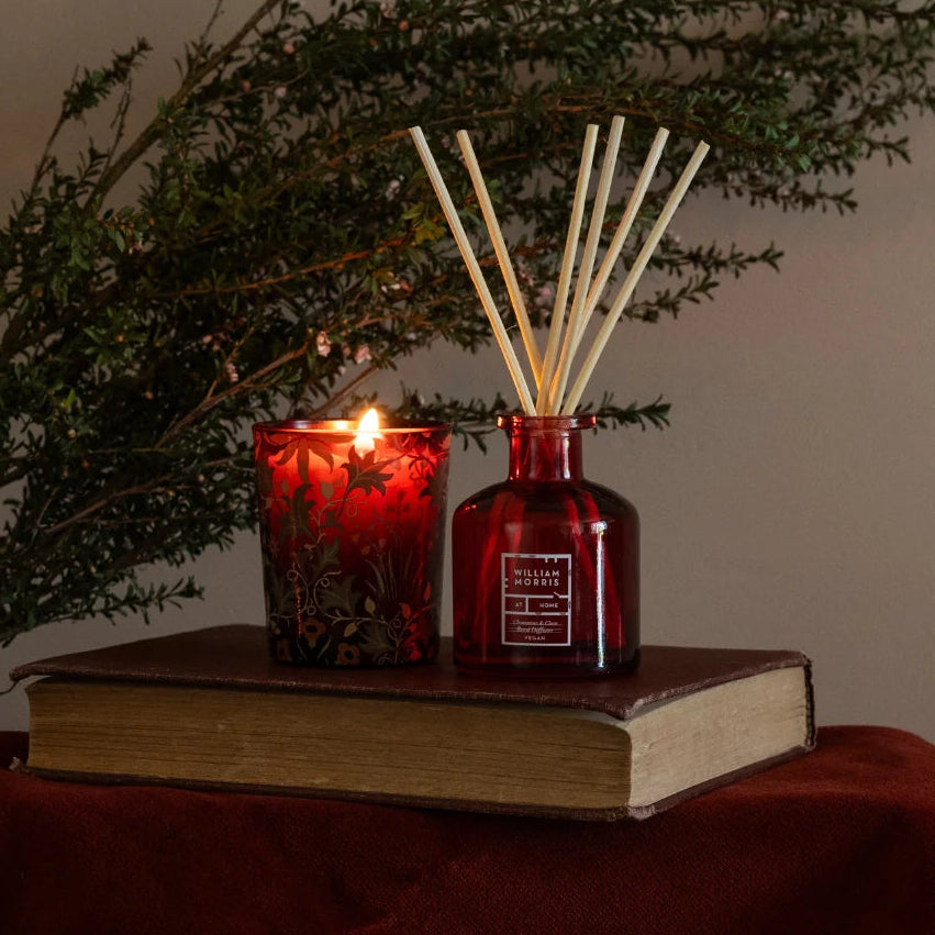 A red glass reed diffuser bottle with wooden reeds and a matching red glass candle holder, styled on top of an old book draped over a dark red cloth with green foliage in the background.
