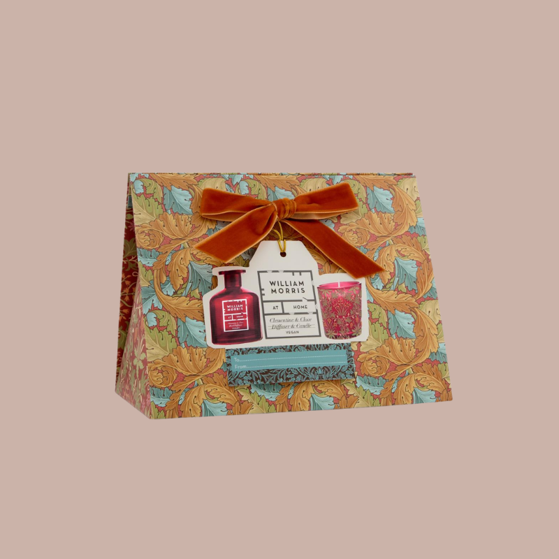 William Morris patterned gift box containing a diffuser and a candle, tied with an orange velvet bow, on a solid beige background.