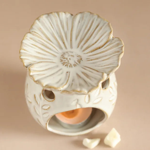 A ceramic flower-shaped candle warmer with a cream-colored glaze and subtle gold-toned edging, featuring a small tea light candle inside and two small white wax pieces in the foreground on a solid beige background 63092 Uneeka