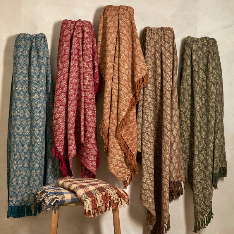 Five woven throw blankets in different earthy colors hang against a light wall, with one plaid blanket draped over a wooden stool. The blankets feature a repeating geometric pattern and fringed edges.